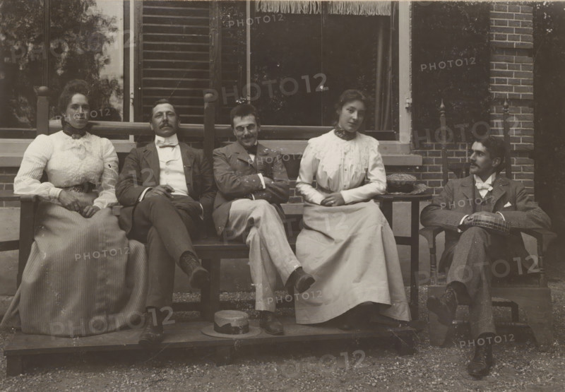 Group portrait with Hedwig Kleintjes, Gerard Aalbersberg, Jan Kleintjes and an unknown man and woman, photograph, Heerde (possibly), c. 1900 - c. 1906, paper, gelatin silver print, developing gelatin silver print, height 0 mm x width 0 mm, Five people sit on wooden chairs in front of a building with a window and brickwork, all wearing formal clothing from an earlier era; two women wear long light-colored dresses with high collars and lace or ruffled details, and three men wear suits with bow ties or neckwear and neatly groomed facial hair. The central man and the man to his right sit with legs crossed and arms folded while the men at the ends sit more relaxed with hands resting on laps or on armrests; a straw hat rests on the wooden platform at their feet and a small round woven basket sits on a side table beside one woman. The background shows shuttered and curtained windows framed by wooden posts and decorative finials on the chair backs, and the ground in front of the platform is gr...