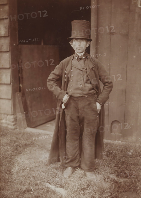 The brother of the corpse', man with top hat in Nunspeet, photograph, title: 'The brother of the corpse Nunspeet', photographer: Gerard Aalbersberg (us), Nunspeet, c. 1897-09, Gerard Aalbersberg (us), photographer, paper, daylight gelatin silver print, height 167 mm x width 118 mm, A sepia-toned photograph of a person standing outdoors in front of a wooden doorway, wearing a tall top hat, a long overcoat, a double-breasted vest with visible buttons, trousers, and shoes, with hands tucked into pockets and a patterned neckerchief at the collar; the background shows vertical wooden boards and a partially open door with a dark interior, and the foreground displays grass underfoot.