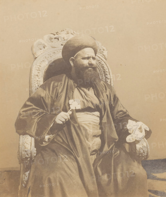 Portrait of a Coptic Patriarch in Cairo, photograph, inscription: '1859 La patriarche des Koptes au Kaire', inscription: '1082, 7 (M v W)', photographer: Willem de Famars Testas (possibly), Cairo (city), 1859, Ottoman Empire, North Africa, paper, paint (coating), deck paint, salted paper print, salt print, white body paint and drawn, height 186 mm x width 159 mm, historical persons not known by name, A seated man wearing a rounded head covering and a long dark robe is shown in a carved ornate chair; he has a full beard and a serious facial expression, one hand resting on the arm of the chair holding a small object and the other hand holding a folded patterned cloth, with a plain light background behind him.