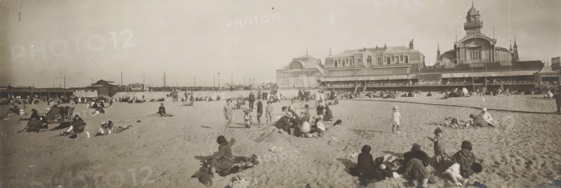 Beach on the North Sea coast, Belgium or France, panoramic photograph, Belgium, France, 1920 - 1940, paper, gelatin silver print, height 134 mm x width 385 mm, beach (as place of recreation), North Sea, A wide sandy beach scene with many people scattered across the sand, some sitting or reclining on blankets and towels, others standing or walking, and a few children playing near small mounds of sand; in the background a long seaside pavilion or complex of ornate buildings with large windows and a tower rises along the shoreline, with a covered veranda where more people are visible; beach equipment such as folding deck chairs and umbrellas appear among the visitors, and distant low structures and fencing line the horizon beneath a clear sky.