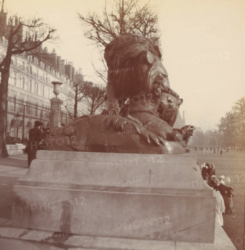 Tuileries, photograph, Paris, c. 1900, photographer, paper, pasteboard, baryta paper, daylight paper print, height 86 mm x width 82 mm, height 92 mm x width 90 mm, piece of sculpture, reproduction of a piece of sculpture, public gardens, park, A large bronze lion sculpture atop a stone pedestal fills the foreground, with the lion's head and mane visible in profile and one paw resting on another animal figure; behind the sculpture a row of leafless trees lines a paved promenade and an iron fence runs along the left side in front of tall multi-story buildings with chimneys; several people in period clothing and hats stand or walk near the fence and beside the pedestal, and a small group of figures is visible further down the promenade; the sky is light and mostly clear.
