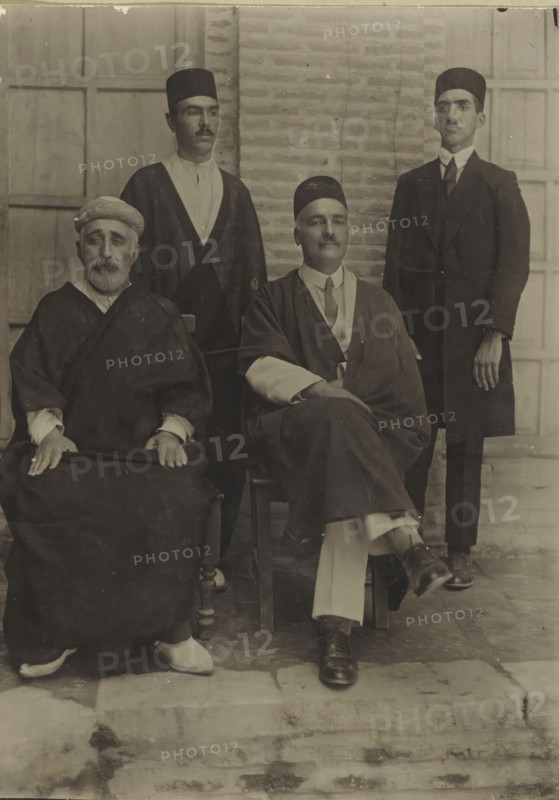 Portrait of Tavakoli (right) and 3 parliamentarians or ministers, photograph, Iran, 1900 - 1950, photographer, gelatin silver print, developing gelatin silver print, height 175 mm x width 127 mm, A sepia-toned portrait shows four men arranged in two rows against a brick and paneled background; two men sit in front on wooden chairs while two men stand behind them. The seated man on the left has a beard, wears a wrapped head covering and a dark robe with visible sleeve cuffs, and rests his hands on his lap where a dark fabric covers his knees; light-colored shoes peek out below his robe. The seated man on the right wears a dark brimless cap, a layered outfit with a lighter shirt and a tie, a draped outer garment, and has one leg crossed over the other with polished dark shoes. Behind them, the standing man on the left wears a brimless cap, a light shirt with a dark outer robe, and has a thin mustache. The standing man on the right wears a brimless cap and a dark suit with a high-collared...
