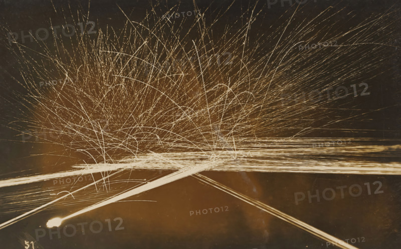 Barrage with machine gun(s) at evening or night, photograph, name: 'PHOTO WIDMER, WALLENSTADT', inscription: 'Tir de barrage, mitrailleuse, FM -, FJ', number: '51', photographer: E. Widmer, c. 1930, paper, gelatin silver print, developing gelatin silver print, height 88 mm x width 139 mm, A dark background filled with numerous thin, bright streaks radiating outward and upward from a lower central area, creating arcs and straight lines that intersect and overlap; a dense cluster of fine, light trails near the center spreads into longer, sparser streaks toward the edges; a few thicker, brighter linear bands run diagonally across the lower portion of the image, one ending in a small bright blob at the left side; overall tones range from warm brown to pale cream for the light trails against a deep, muted backdrop.