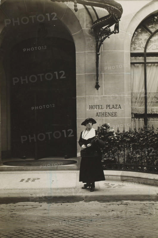 Woman on the street with a bouquet, photograph, inscription: 'Àffaire Costa', Paris, 1916, photographer, paper, gelatin silver print, developing gelatin silver print, height 117 mm x width 80 mm, A monochrome photograph of a woman standing on a tiled entrance in front of a stone building with arched openings; she wears a dark long skirt and jacket with a contrasting light collar and a wide-brimmed hat, holds a small handbag and a large bouquet of flowers, and stands beside an ornate iron railing covered with foliage; the building façade shows a decorative metal canopy and a curtained window with patterned fabric, and the left side of the image reveals a dark recessed archway leading inward.