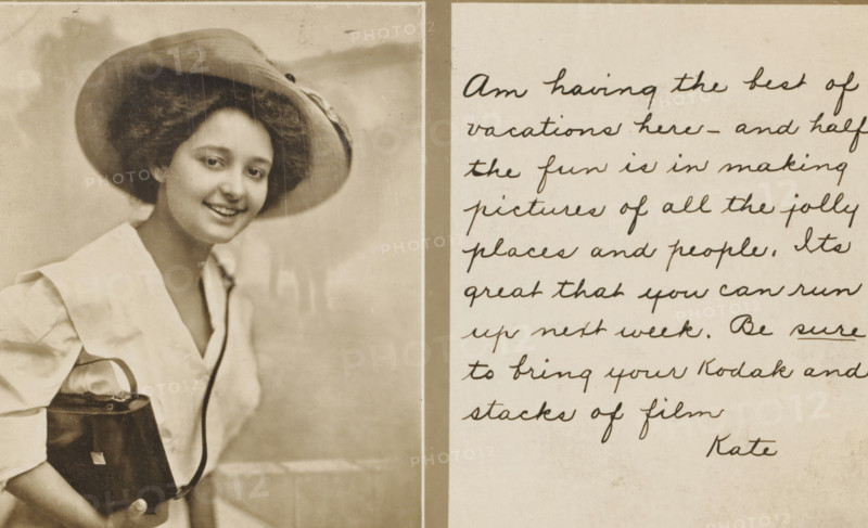 Advertisement for Kodak, picture postcard, stamp: '1910', inscription, inscription: 'Am having the best of, vacations here – and half, the fun is in making, pictures of all the jolly, places and people. Its, great that you can run, up next week. Be sure, to bring your Kodak and, stacks of film, Kate', United States of America, printer (person): Eastman Kodak, in or before 1910, photographer, printer (person), North America, paper, gelatin silver print, developing gelatin silver print, height 150 mm x width 100 mm, Person, A sepia-toned photograph shows a smiling young woman wearing a wide-brimmed hat and a light-colored blouse with a dark trim at the neckline, holding a dark rectangular handbag with a handle and strap, positioned beside a vertical pale border that separates her portrait from a lighter panel on the right that contains rows of cursive handwriting.