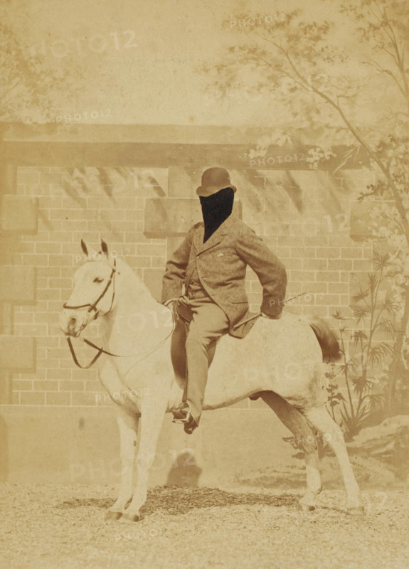 Man on horseback with his face retouched, photograph, United States of America, c. 1880 - c. 1910, photographer, North America, paper, pasteboard, albumen print, albumen print on cardboard, height 140 mm x width 102 mm, height 157 mm x width 119 mm, horse, A sepia-toned photograph of a person seated on a light-colored horse in front of a brick wall with a horizontal ledge and some foliage at the top and right side; the person wears a bowler-style hat and a textured jacket with trousers and has one hand resting on the horse's back while the other holds the reins, and the horse wears a bridle and stands with its head turned slightly toward the left side of the image.