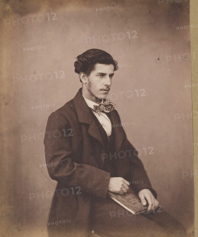 Portrait of an unknown seated young man with a checkered bow tie, with a book in his hand, photograph, photographer: attributed to Franz Hanfstaengl, Germany, c. 1855 - c. 1860, Attributed to, paper, salted paper print, salt print, height 189 mm x width 154 mm, height 238 mm x width 239 mm, anonymous historical person portrayed, A young man is seated against a plain background, shown from the waist up and turned slightly to his right; he has dark, wavy hair styled away from his face and faint sideburns, a straight nose, and a neutral expression with his gaze directed off to the right. He wears a dark coat over a fitted vest and a light-colored shirt with a patterned bow tied at the collar. One hand rests on his lap while the other holds a rectangular object with a patterned cover. The photograph has a sepia tone and shows slight aging marks and small spots on the background and edges.