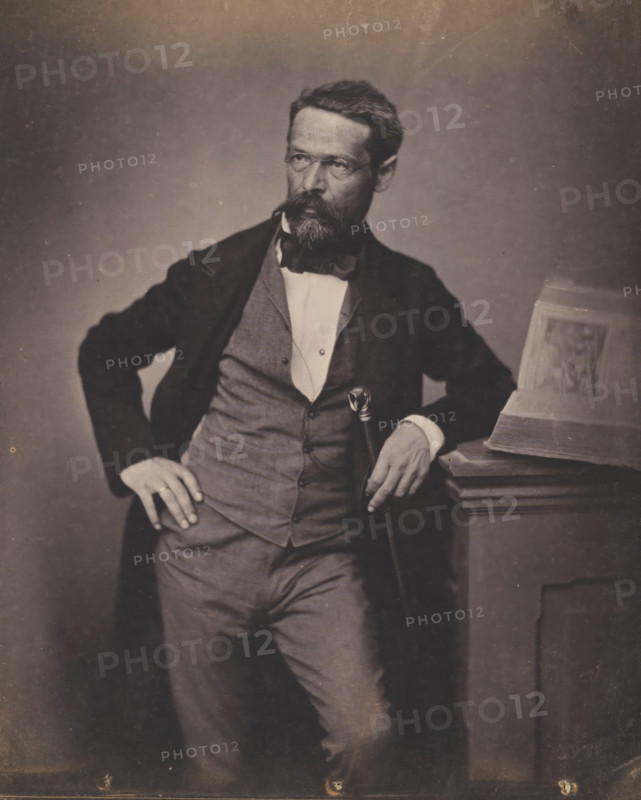 Portrait of an unknown man with glasses and beard, photograph, inscription: 'Ingammikonz', photographer: attributed to Franz Hanfstaengl, Germany, c. 1855 - c. 1860, Attributed to, paper, salted paper print, salt print, height 205 mm x width 154 mm, height 238 mm x width 239 mm, anonymous historical person depicted, A seated man in formal attire with a beard and mustache leans against a wooden pedestal while holding a cane, wearing a dark jacket over a waistcoat and a white shirt with a bow tie, one hand resting on his hip and the other on the pedestal where an open book is placed, the background is plain and the image has a muted, sepia tone.