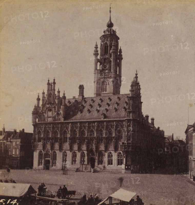 View of the town hall of Middelburg, Town Hall in Middelburg, Views and landscapes in Europe, Africa and Asia, Geographical Sculpture Gallery, collected by MJ Broekhuijsen, Headmaster of a school in Amsterdam, stereograph, Numbered lower left of photo: F.14., Middelburg, publisher: Merkelbach & Co., Amsterdam, compiler (person in literature): Martien Johan Broekhuijsen, c. 1905, photographer, compiler (person in literature), pasteboard, paper, albumen print, height 88 mm x width 178 mm, Part of a group of 52 stereophotos used in primary education as a supplement to school charts. The entire series consisted of 274 numbers in 10 categories (A to K)., town hall, A large ornate stone building with Gothic architectural details occupies the center, featuring a steep roof punctuated by dormer windows and decorative pinnacles, a tall central tower with an onion-shaped spire and smaller turrets, and a facade of arched windows and sculpted stonework; in front of the building there is an open pa...