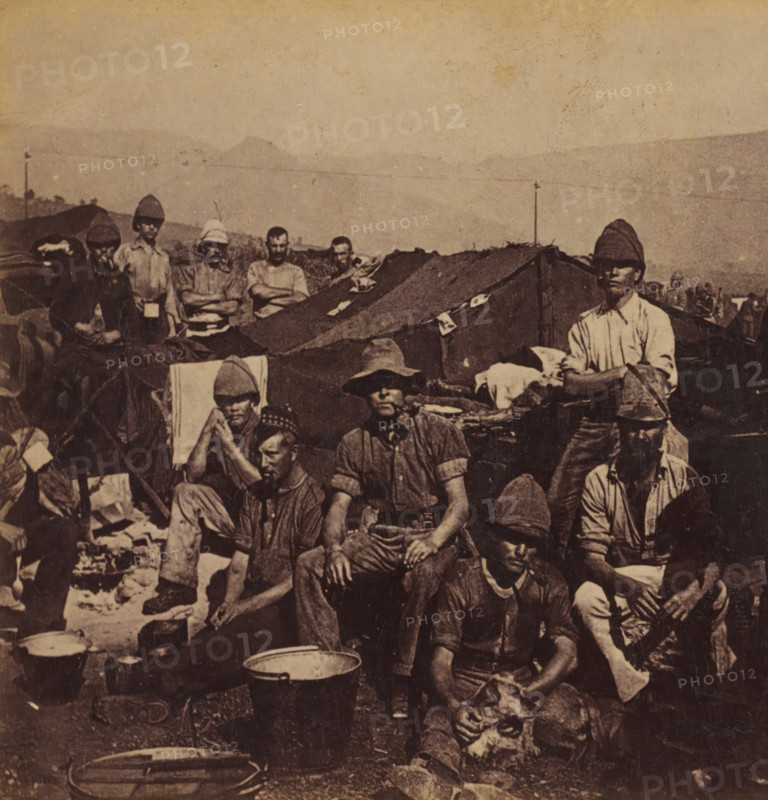 Cold Stream guards in their camp, Guards on watch in South Africa during the Second Boer War, Faces and landscapes in Europe, Africa and Asia, Geographical Sculpture Gallery, collected by MJ Broekhuijsen, Headmaster of a school in Amsterdam, stereograph, annotation: 'See pl. 9.', South Africa, publisher: Merkelbach & Co., Amsterdam, compiler (person in literature): Martien Johan Broekhuijsen, c. 1899 - c. 1905, photographer, compiler (person in literature), pasteboard, paper, albumen print, height 86 mm x width 177 mm, Part of a group of 52 stereophotos used in primary education as a supplement to school charts. The entire series consisted of 274 numbers in 10 categories (A to K)., tent-camp, guard duty, keeping sentry, A group of men wearing varied hats and casual shirts are gathered outdoors among canvas tents, blankets, and scattered personal items; some men sit while others stand with arms crossed or resting on knees, and several faces show neutral expressions; a large cooking pot ...
