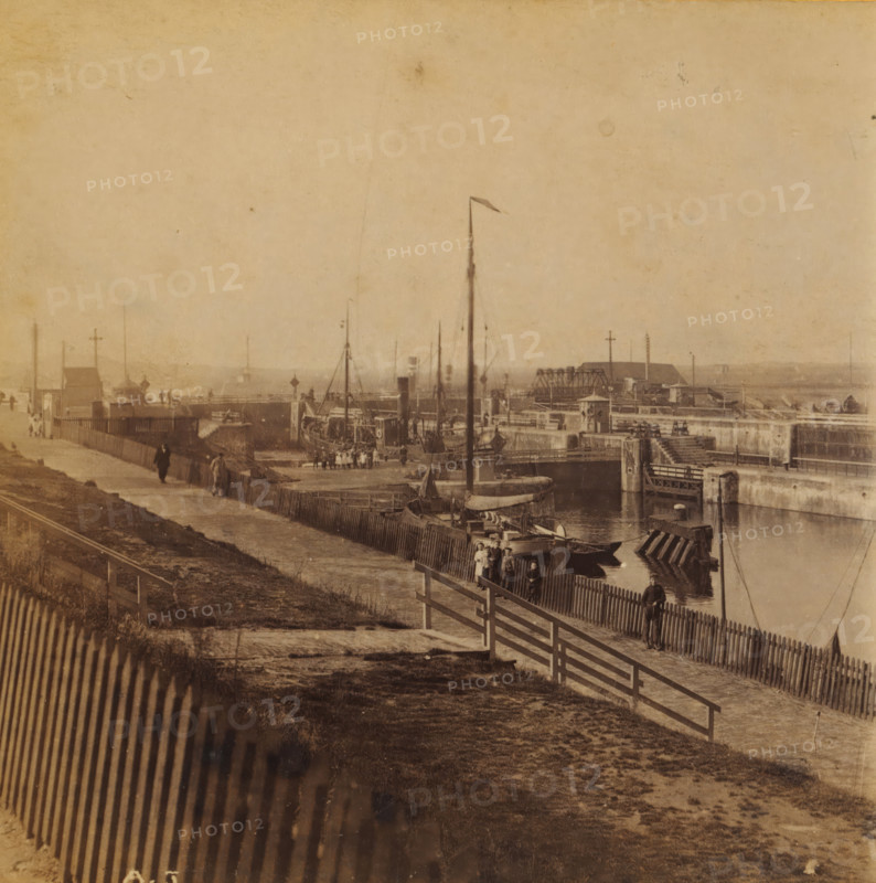IJmuiden, View of IJmuiden southeast of the harbor, Views and landscapes in Europe, Africa and Asia, Geographical Sculpture Gallery, collected by MJ Broekhuijsen, Headmaster of a school in Amsterdam, stereograph, Numbered lower left of photo: A.5., annotation: 'We are standing southeast of the harbor. A boat is currently being locked through the lock.', publisher: Merkelbach & Co., Amsterdam, compiler (person in literature): Martien Johan Broekhuijsen, c. 1905, photographer, compiler (person in literature), pasteboard, paper, albumen print, height 86 mm x width 176 mm, Part of a group of 52 stereophotos used in primary education as a supplement to school charts. The entire series consisted of 274 numbers in 10 categories (A to K)., city-view in general, 'veduta', traffic on the water, A sepia-toned waterfront scene with a fenced pathway running alongside a harbor area where several small boats and larger vessels are moored; wooden piers, posts, and a tall mast with a small flag rise fr...