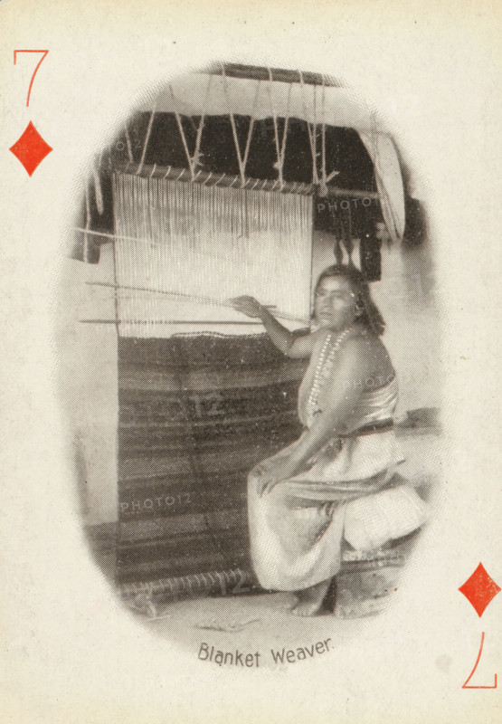 Blanket Weaver, Seven of Diamonds with woman at loom weaving a blanket, photograph, playing card, Playing card from a box of 53 playing cards with autotype images of Native Americans. The contents include a complete set of playing cards including a joker., photographer: Adam Clark Vroman, United States of America, publisher: Lazarus & Melzer, Los Angeles (city), c. 1900, North America, paper, autotype, height 88 mm x width 63 mm, A seated person wearing a draped garment and a beaded necklace is positioned beside a large vertical loom with threads hanging and woven fabric visible, the person’s arm is extended toward the loom and they are looking toward the viewer, the scene appears within an oval vignette on a textured light background with a red diamond shape near the top left and another near the lower right.