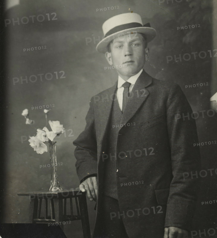Rapid photographs, Snelfoto, photographer: Jan Gottfried Stütner, Alkmaar, 1913-07-09 - 1926, paper, gelatin silver print, developing gelatin silver print, height 66 mm x width 60 mm, A black and white portrait of a man wearing a three-piece suit and a boater hat with a dark band, standing beside a small wooden table on which a tall glass vase holds several light-colored flowers, the man’s hand resting on the table edge and his other hand relaxed at his side, with a plain, slightly mottled backdrop and some visible surface scratches on the photograph.