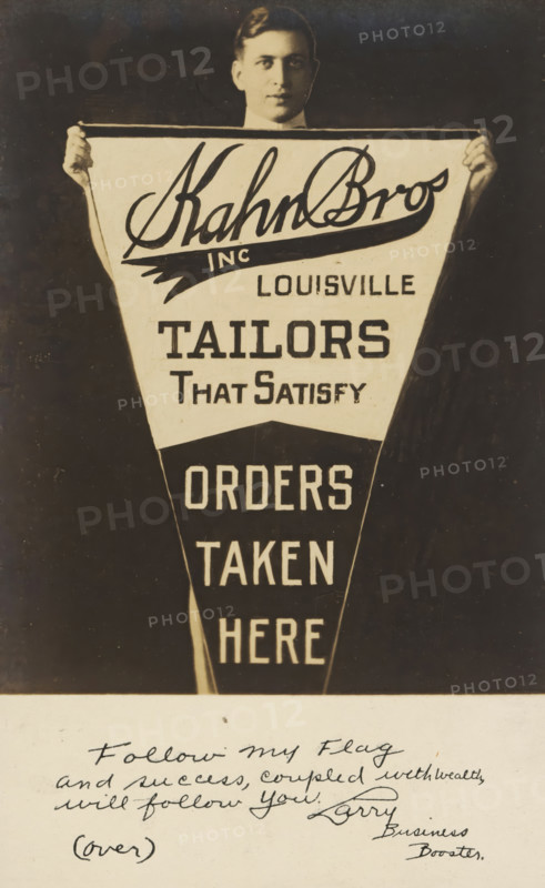 Advertisement for Kahn Bros, Louisville, picture postcard, inscription, United States of America, c. 1930 - c. 1950, photographer, North America, paper, gelatin silver print, developing gelatin silver print, height 138 mm x width 89 mm, Person, Kahn Bros, Kentucky, A sepia-toned photograph of a man seen from the chest up holding a large triangular pennant in front of him, his hands gripping the top corners and his face partially visible above the pennant with short dark hair and a neutral expression; the pennant has alternating light and dark sections with bold graphic lettering and blocky shapes, and there is a lighter rectangular strip across the top edge; below the pennant the image shows a lighter paper area with cursive handwriting and a small parenthetical word, the handwriting slants and fills the space with several lines of flowing script.