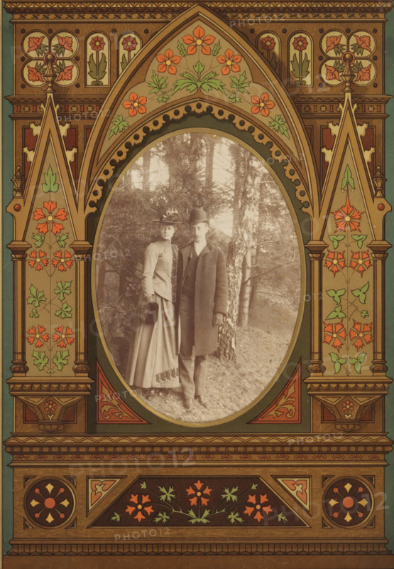 Portrait of a man and a woman in a forest, album page, photograph, cabinet photograph, This photo is part of an album., 1850 - 1900, photographer, paper, pasteboard, albumen print, height 136 mm x width 96 mm, historical persons, historical persons - BB - woman, forest, wood, A vertically oriented ornate frame with a pointed arch motif surrounds an oval photograph; the frame is decorated with stylized floral patterns in muted reds, greens, and gold tones, with symmetrical panels, columns, and carved-looking details; inside the oval, a sepia-toned portrait shows a man and a woman standing outdoors on grass in front of trees, the woman wearing a long skirt, fitted jacket, and a hat with a decorative element while holding a muff, and the man wearing a long overcoat, vest, trousers, and a bowler-style hat, both facing the camera and positioned close together.