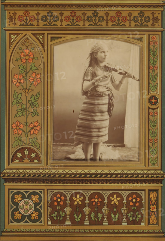 Portrait of a girl playing the violin, album page, photograph, cabinet photograph, This photo is part of an album., 1850 - 1900, photographer, paper, pasteboard, albumen print, height 136 mm x width 96 mm, historical persons - BB - woman, violin, fiddle, girl (child between toddler and youth), A sepia photograph of a young girl standing and playing a violin is centered within a decorative, floral border. She wears a striped dress with a sash tied at her waist and a patterned headscarf draped over her hair, and bracelets on her wrists. The girl looks slightly upward and holds the violin under her chin with one hand and the bow with the other. The background behind her is plain and light, with a curtain visible at one side and a fur rug at her feet. The surrounding frame features symmetrical ornamental panels with stylized flowers, leaves, geometric motifs, and warm earthy colors, including shades of brown, green, and orange, forming arch shapes and repeating floral patterns.