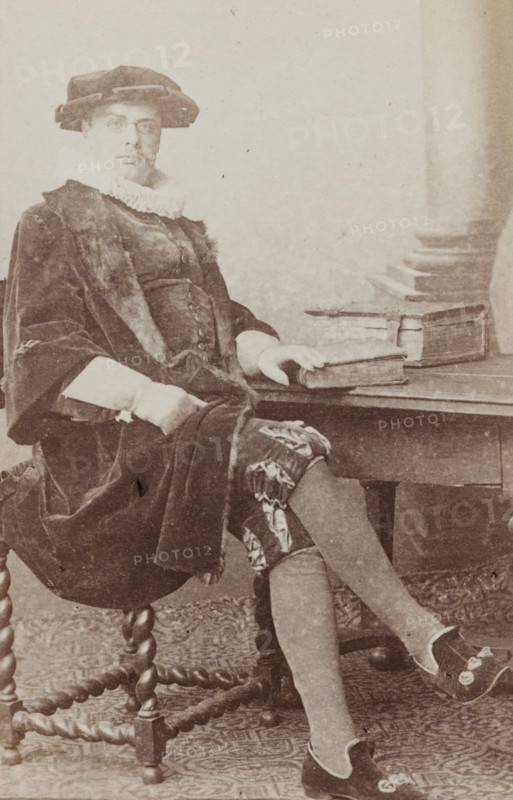 Portrait of a man in a suit sitting at a table, identified as N. Dirkzwager, album page, photograph, carte-de-visite, This photo is part of an album., name: 'N. Dirkzwager. 49', inscription: 'J. Goedeljee, Photographer, HRH PRINCE ALEXANDER, of the Netherlands, PRINCE of ORANGE, and HRH, PRINCESS HENDRIK, Hoogewoerd 160, near the Plantage.', photographer: Jan Goedeljee, Leiden, 1860 - 1905, paper, pasteboard, albumen print, height 83 mm x width 52 mm, historical persons, table, historical costume, A sepia portrait of a seated person wearing a dark velvet coat with fur trim and a ruffled white collar, a wide brimmed hat, patterned knee breeches, and decorative slippers, sitting on a carved wooden chair with twisted legs and resting one hand on a stack of books placed on a wooden table beside them.