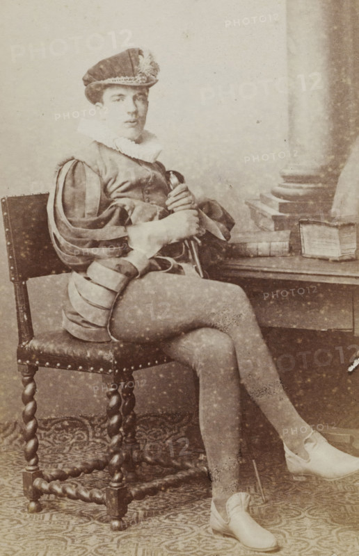 Portrait of a man in a suit sitting at a table with books, identified as J. Wiarda, album page, photograph, carte-de-visite, This photo is part of an album., inscription: 'J. Goedeljee, Photographer, HRH PRINCE ALEXANDER, of the Netherlands, PRINCE of ORANGE, and HRH, PRINCESS HENDRIK, Hoogewoerd 160, near the Plantage.', name: 'J. Wiarda, 52.', photographer: Jan Goedeljee, Leiden, 1860 - 1905, paper, pasteboard, albumen print, height 83 mm x width 52 mm, historical persons, historical costume, A seated young person in a historical costume with a brimmed hat adorned with trim and a ruffled collar looks toward the camera. They wear a doublet with slashed sleeves revealing inner fabric and fitted hose, and hold a slender object across their lap with both hands. The chair has a studded leather seat and twisted wooden legs, and a table beside them holds a closed book and an open book resting on its side. A patterned floor covering extends beneath the chair and their feet, and a column deta...