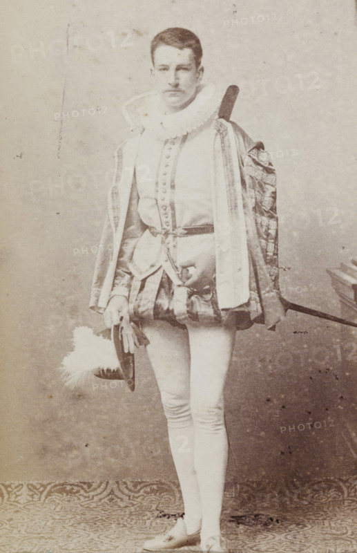 Portrait of Tj.AMA Humalda van Eysinga in costume with hat in hand, album page, photograph, carte-de-visite, This photo is part of an album., inscription: 'J. Goedeljee, Photographer, HRH PRINCE ALEXANDER, of the Netherlands, PRINCE of ORANGE, and HRH, PRINCESS HENDRIK, Hoogewoerd 160, near the Plantage.', name: 'Tj. AMA Humalda, van Eysinga., 93.', photographer: Jan Goedeljee, Leiden, 1860 - 1905, paper, pasteboard, albumen print, albumendruk, height 83 mm x width 52 mm, Person, Tj.AMA Humalda van Eysinga, historical persons, head-gear: top hat, historical costume, A full-length sepia photograph of a person standing in ornate historical costume, wearing a ruffled collar and a decorated robe with patterned trim draped over one shoulder, a buttoned doublet with a belted waist and a short padded skirt, fitted hose, and flat shoes; the person holds a wide-brimmed hat with a large plume in one hand and a sword hangs at the waist and over the shoulder; the background is plain and the floor ...