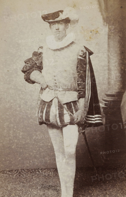Portrait of a young man in costume with a sword, identified as JAE Rhede van der Kloot, album page, photograph, carte-de-visite, This photo is part of an album., name: 'J. AE. Rhede vd Kloot., 100.', inscription: 'J. Goedeljee, Photographer, HRH PRINCE ALEXANDER, of the Netherlands, PRINCE of ORANGE, and HRH, PRINCESS HENDRIK, Hoogewoerd 160, near the Plantage.', photographer: Jan Goedeljee, Leiden, 1860 - 1905, paper, pasteboard, albumen print, height 83 mm x width 52 mm, historical persons, historical costume, hacking and thrusting weapons: rapier, A faded sepia photograph of a standing person wearing a historical costume with a plumed hat, a ruffled collar, a doublet with decorative buttons, puffed sleeves, a short striped skirt-like garment, fitted hose, and a cape draped over one shoulder; the person has one hand resting on their hip and the other holding a sword at their side; a column and draped backdrop are visible to one side and a patterned floor covering is beneath their fee...