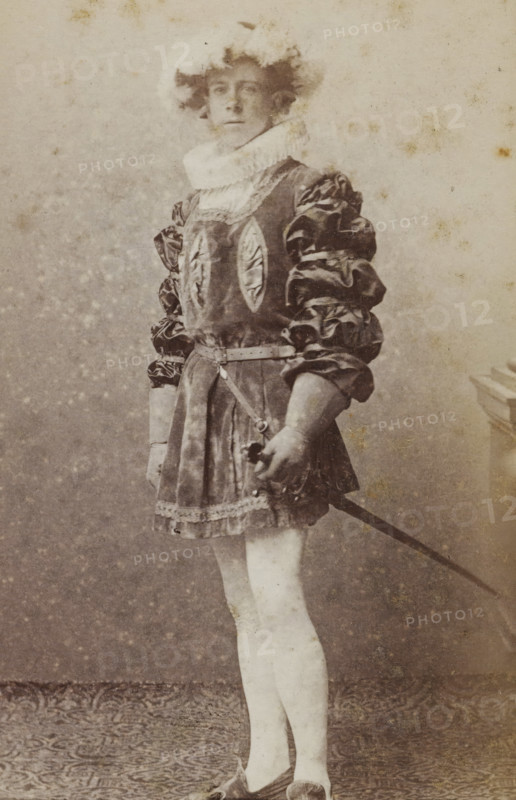 Portrait of Jan Gerritzen in costume with sword, album page, photograph, carte-de-visite, This photo is part of an album., name: 'J. Gerritzen., 102.', inscription: 'J. Goedeljee, Photographer, HRH PRINCE ALEXANDER, of the Netherlands, PRINCE of ORANGE, and HRH, PRINCESS HENDRIK, Hoogewoerd 160, near the Plantage.', photographer: Jan Goedeljee, Leiden, 1886 - 1892, paper, pasteboard, albumen print, albumendruk, height 83 mm x width 52 mm, Person, Jan Gerritzen, historical persons, historical costume, hacking and thrusting weapons: rapier, A faded sepia photograph of a person standing in a full-length pose wearing a costume with a short skirt and puffed sleeves, a ruffled high collar around the neck, and a belt with a strap crossing the waist; the left hand holds the hilt of a sword whose blade extends downward and out of frame, while the right hand rests near the hip; the person has light-colored stockings and pointed shoes, and a hat or headpiece with fluffy details sits atop tousled ...