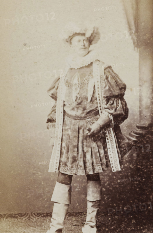 Portrait of a young man in costume, identified as BPG van Diggelen, album page, photograph, carte-de-visite, This photo is part of an album., inscription: 'J. Goedeljee, Photographer, HRH PRINCE ALEXANDER of the Netherlands, PRINCE of ORANGE, and HRH PRINCESS HENDRIK of the Netherlands, Hoogewoerd 160, near the Plantage.', name: 'BPG v. Diggelen., 247.', photographer: Jan Goedeljee, Leiden, 1860 - 1905, paper, pasteboard, albumen print, height 83 mm x width 52 mm, historical persons, historical costume, A sepia-toned photograph of a person standing and facing slightly to the side, wearing a historical costume with a ruffled collar and a decorative bodice, puffed sleeves and long ornate ribbons hanging from the shoulders down the front of the outfit, a pleated skirt with lace trim and a patterned apron-like overlay, tall layered boots covering the calves, and a hat with a feather.