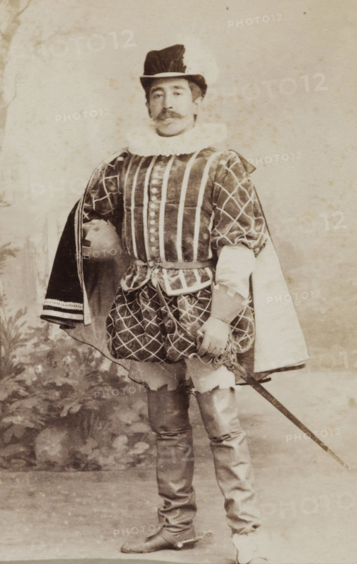 Portrait of a young man in costume with sword, identified as W. Birnie, album page, photograph, carte-de-visite, This photo is part of an album., inscription: 'Ant. J. van der Stok, PHOTOGRAPHE, Leiden Rapenburg 13.', name: 'W. Birnie 249.', photographer: Antonius Joannes van der Stok, Leiden, 1887-01-01 - 1899-12-03, paper, pasteboard, albumen print, albumendruk, height 83 mm x width 52 mm, historical persons, hacking and thrusting weapons: sword, historical costume, A sepia-toned photograph of a man standing and facing forward wearing a ruffled collar and a tall hat with a brim. He has a mustache and is dressed in a patterned doublet with vertical stripes on the front and diamond-patterned sleeves, and a short, quilted skirt over fitted hose. One arm rests on his hip beneath a cloak draped over his shoulder, and the other hand holds the hilt of a sword that extends downward. He wears tall boots with turned-down tops and the background shows a plain, lightly mottled backdrop with fain...