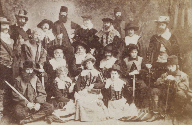 Group portrait of students in costume, album page, photograph, carte-de-visite, This photo is part of an album., inscription: 'J. Goedeljee, Photographer, HRH PRINCE ALEXANDER of the Netherlands, PRINCE of ORANGE, and HRH PRINCESS HENDRIK of the Netherlands, Hoogewoerd 160, near the Plantage.', photographer: Jan Goedeljee, Leiden, 1880 - 1900, paper, pasteboard, albumen print, height 83 mm x width 52 mm, student, anonymous historical persons portrayed in a group, in a group-portrait, historical costume, A sepia photograph showing a large group of people arranged in rows, many wearing period costumes with wide collars, hats, cloaks, and beards; some individuals hold swords and one person rests a hand on a cane; central figures include seated women in striped bodices and full skirts, surrounded by men in varied headwear from wide-brimmed hats to fur caps and tall hats, and a few figures wear fur-trimmed garments; expressions are mostly neutral, and the background is plain and softly text...
