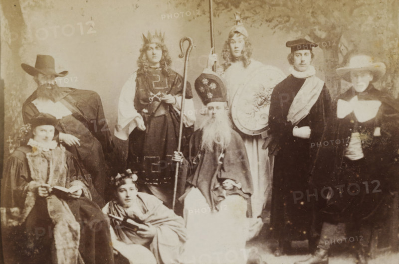 Group portrait of students in costume, album page, photograph, carte-de-visite, This photo is part of an album., inscription: 'J. Goedeljee, Photographer, HRH PRINCE ALEXANDER of the Netherlands, PRINCE of ORANGE, and HRH PRINCESS HENDRIK of the Netherlands, Hoogewoerd 160, near the Plantage.', photographer: Jan Goedeljee, Leiden, 1880 - 1900, paper, pasteboard, albumen print, height 83 mm x width 52 mm, student, anonymous historical persons portrayed in a group, in a group-portrait, historical costume, A sepia-toned group portrait showing several people in period or theatrical costumes arranged in two rows, with some seated and others standing; visible are ornate hats and headpieces including a crown, a tall mitre-like hat, and wide-brimmed hats, along with long robes, capes, fur-trimmed garments, and a variety of collars and sashes; one seated figure holds a staff and has a long beard, another standing figure holds a tall pole, a seated person holds an open book, and a reclining pers...
