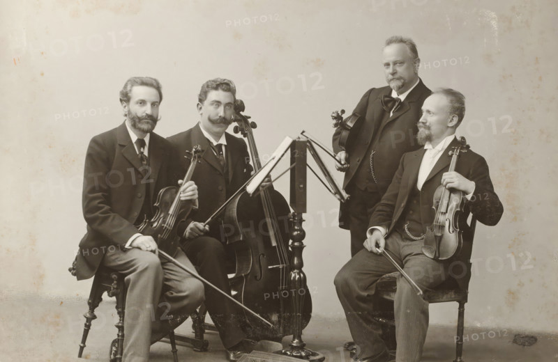 Group portrait of a string quartet, album page, photograph, cabinet photograph, This photo is part of an album., photographer: Wilhelm Höffert, 1901, paper, pasteboard, gelatin silver print, developing gelatin silver print, height 134 mm x width 196 mm, historical persons, adult man, small group of musicians, chamber orchestra, jazz band, A black-and-white photograph of four seated men in formal suits arranged around a central music stand, each holding a string instrument; two men hold violins with bows visible, one man holds a cello with its endpin on the floor, and one man holds a viola while resting a bow on his knee. The men have varied facial hair styles including mustaches and beards, and they wear dark jackets, waistcoats, and ties or cravats. The music stand has an open score visible on its ledge and slender metal supports. The chairs are wooden with turned legs and carved details. The background is plain and light-colored with some mottled spots and age marks on the image surf...