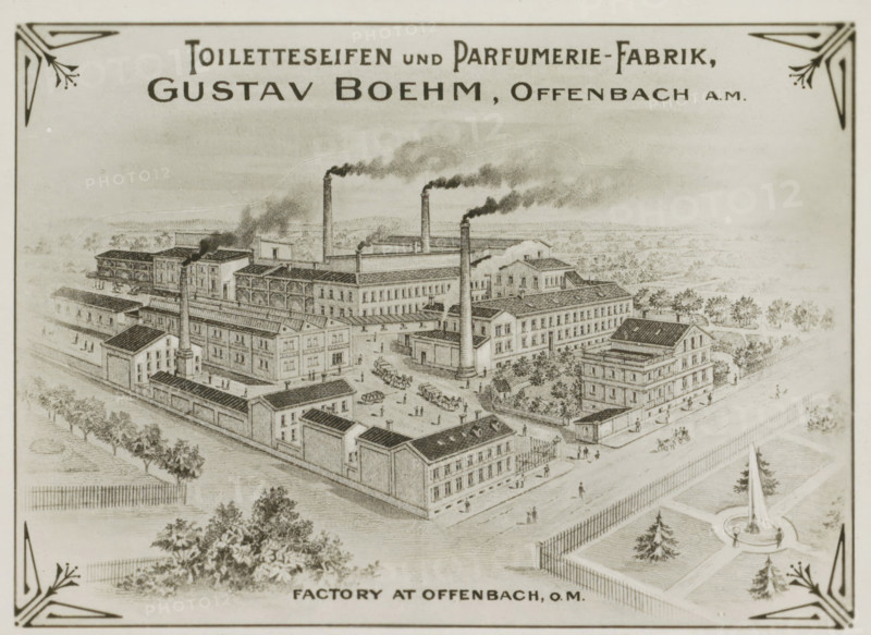 Photo reproduction of (presumably) a print of Gustav Boehm's soap and perfume factory in Offenbach am Main, photograph, reproduction, Part of Photographs of Gustav Boehm's world tour 1899-1901, series 2., after print by anonymous, 1899 - 1901, photographer, after print by, paper, gelatin silver print, developed gelatin silver print, height 82 mm x width 60 mm, factory-building, Hessen, Offenbach am Main, A sepia-toned engraved illustration of an industrial complex seen from an elevated perspective, showing multiple rectangular brick buildings with pitched and flat roofs arranged around courtyards and streets, tall smokestacks emitting plumes of smoke, small figures of people walking and standing, horse-drawn carts and carriages on the roads, rows of trees and fenced garden areas surrounding the factory, a circular fountain feature with a tall central obelisk in a landscaped area, and decorative corner motifs framing the image.