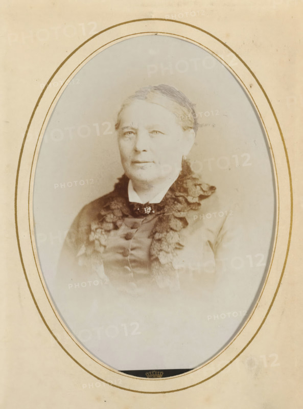 Portrait of a woman, album page, photograph, cabinet photograph, This photo is part of an album., inscription: ', Dupont, 67, Rue Neuve, Bruxelles, ', photographer: Dupont (photo studio), 1864 - 1886, paper, pasteboard, albumen print, height 134 mm x width 96 mm, historical persons, adult woman, An oval-framed sepia portrait of an older woman shown from the chest up, wearing a high-collared dress with a dark ruffled trim and a small decorative brooch at the throat, her hair pulled back and covered with a thin headband, her expression neutral, set against a pale, slightly mottled background and surrounded by a cream mat with concentric gold lines.