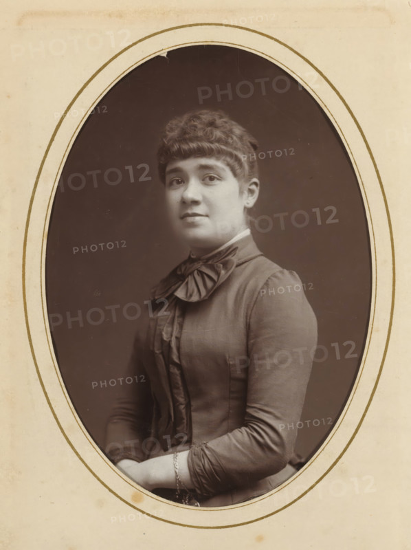 Portrait of a young woman, album page, photograph, cabinet photograph, This photo is part of an album., inscription: 'Inalterable Process, Charcoal Photography, Nestor Schaffers, Photographer, Rue Guinard 9, Ghent', inscription: 'Inalterable charcoal impression, photographer: Paul Joseph Nicolas Nestor Schaffers, 1875 - 1896, Paul Joseph Nicolas Nestor Schaffers, photographer, paper, pasteboard, carbon print, height 134 mm x width 96 mm, historical persons, adolescent, young woman, maiden, A sepia portrait of a young woman shown from the waist up inside an oval frame, wearing a high-collared dress with a large bow at the neck and decorative vertical ruffles down the front, her hair styled with short bangs and a curled updo, she faces slightly toward the camera with a neutral expression, her hands folded in her lap with a delicate bracelet visible on one wrist, the background inside the oval is plain and darker than the surrounding light-colored mat.