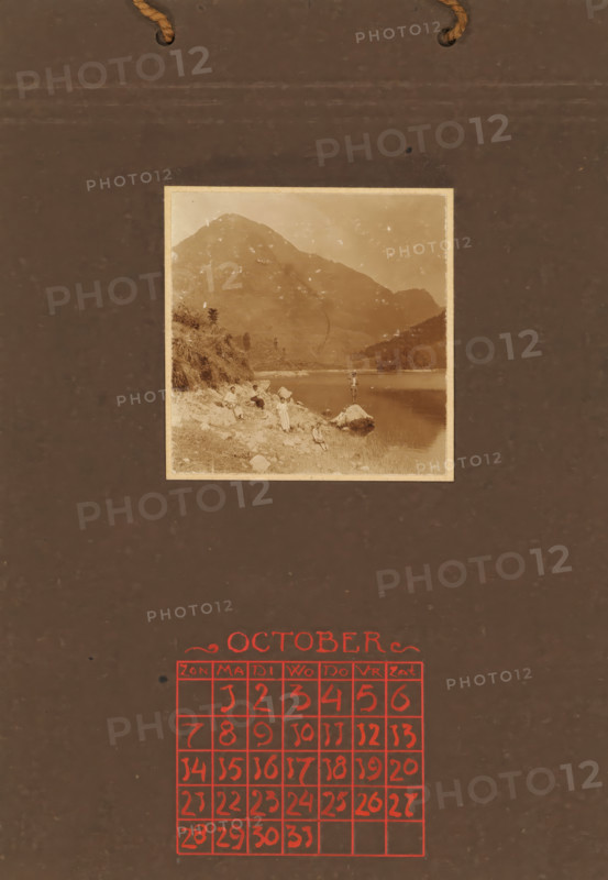 Three women, a man and two children on rocks on the coast, with a mountain in the background, album page, photograph, This photo is part of an album., annotation: 'OCTOBER, ', Indonesia, 1880 - in or before 1923, photographer, pasteboard, paper, daylight collodion silver print, height 80 mm x width 78 mm, adult man, mountains, rocky coast, child (two persons), adult woman (three persons), A sepia-toned photograph mounted on a dark brown textured background shows a lakeshore scene with mountains rising behind the water; the shoreline is rocky with grass and scattered stones, and several small figures in period clothing are positioned along the bank and on a large rock at the water's edge. The background surface has two short pieces of rope threaded through holes near the top and faint scuff marks and spots across the brown area. Below the photograph a grid of hand-drawn squares filled with hand-drawn numerals is printed in a contrasting color.