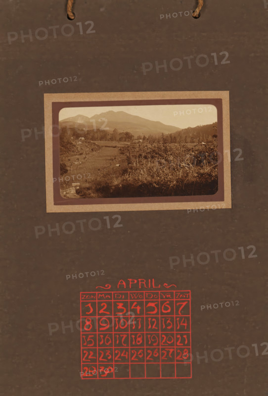 Landscape with rice fields, album page, photograph, This photo is part of an album., annotation: 'APRIL, ', Indonesia, 1880 - in or before 1923, photographer, pasteboard, paper, daylight collodion silver print, height 58 mm x width 101 mm, landscapes, sawahs, rice paddies, A dark brown textured background with two short pieces of rope at the top, a sepia-toned framed photograph of a hilly landscape centered near the upper middle showing layered hills, trees, vegetation, and grassy foreground within a rectangular matte, and a grid of hand-drawn red squares near the lower center forming a calendar layout with a decorative red heading above the grid.