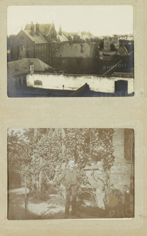 View of a walled prison complex with church in Oslebshausen, photograph, postcard, Part of Photo album of Carl Heinrich Fliegenschmidt, prison director in Oslebshausen., Oslebshausen, 1890 - 1910, photographer, Bremen (city), paper, gelatin silver print, developed gelatin silver print, height 78 mm x width 110 mm, fence, wall, paling, church (exterior), prison, jail, A faded photograph showing an elevated view of a cluster of older brick buildings with steeply pitched roofs and tall chimneys, a high pale wall with a gated opening in the foreground, a darker roofed structure partially obscured by trees behind the wall, and additional rooftops and tree-lined areas receding into the distance.