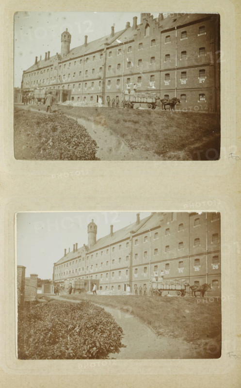 Exterior of Oslebshausen prison with workers by carts, photograph, Part of Photo album of Carl Heinrich Fliegenschmidt, prison director at Oslebshausen., Oslebshausen, c. 1890 - c. 1910, photographer, Bremen (city), pasteboard, paper, daylight collodion silver print, height 78 mm x width 110 mm, working class, laborers, prison, jail, animal traction, animal-drawn vehicle (carriage etc.), A sepia-toned photograph shows a long brick building with many small barred windows and a pitched roof with chimneys and a distinctive round tower rising above the roofline; in front of the building a dirt path curves beside a low hedge and an open grassy area; a horse-drawn wagon loaded with long bundled items stands near the building with a group of people nearby, some standing on the ground and some on the wagon; additional figures and a cart are visible further along the frontage near an arched entrance, and a small piled heap of material lies against the wall.