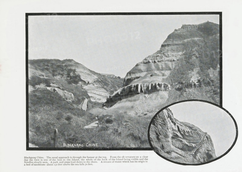 Blackgang Chine, photograph, page, Page 20 with 2 photos of Blackgang Chine and a caption. Part of Photographic travelogue of the Isle of Wight., photographer: Welch & Sons, J., Isle of Wight, in or after 1897 - c. 1930, printer (person), paper, printing, autotype, page with photos and printed text, height 169 mm x width 228 mm, travelling, tourism, ravine, chasm, abyss, canyon, inland cliff face (landscape with figures, staffage), Blackgang, A black and white framed photograph of a coastal valley with layered rock cliffs and steep slopes, showing a narrow winding path and a series of steps descending through grassy and scrubby terrain, scattered vegetation on the hillside, a small structure visible near the middle distance between the cliffs, striated rock formations and ledges on the larger cliff to the right, and an overlapping circular inset at the lower right showing a close-up of a rugged cliff face with a path or railing along its edge.