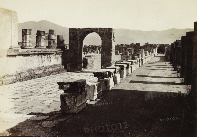 Foro e Tempio di Giove di Pompei., Remains of the forum and the Temple of Jupiter in Pompeii, album page, photograph, Part of Photo album with recordings of excavations in Pompeii by Roberto Rive., number: '407', photographer: attributed to Roberto Rive, Pompeii, c. 1860 - c. 1900, Attributed to, paper, albumen print, height 104 mm x width 141 mm, square, circus, temple, shrine, Roman religion, excavation, archaeology, A wide, sunlit scene of ancient stone ruins with a paved foreground and a long shadowed pathway running alongside a row of low rectangular stone blocks, a prominent arched stone gateway standing near the center, fluted column fragments on a raised platform to the left, scattered carved stone pieces leading toward distant ruined walls and structures, and low hills visible on the horizon under a pale sky.