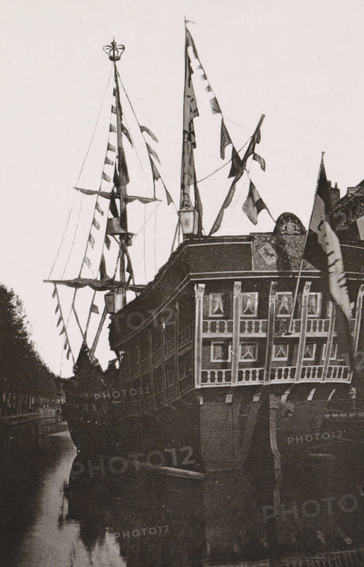 Galley on the Herengracht., Replica of a galley on the Herengracht during the April Festivals in 1887 in Amsterdam, photograph, cabinet photograph, Part of Photo album with images of the April Festivals in 1887 in Amsterdam., Amsterdam, 1887-04, photographer, pasteboard, ink, materials by function, paper, printing ink, collotype, photogravure, height 147 mm x width 97 mm, sailing-ship, sailing-boat, canal, prospect of city, town panorama, silhouette of city, April Festivals 1887, Herengracht (Amsterdam), A large ornate sailing ship is shown from the stern, with tall masts and rigging decorated with multiple triangular and rectangular flags; the ship’s stern has a multi-windowed gallery with curtained panes and decorative railings, and a carved emblem above the gallery. The hull is dark and weathered, and the ship is moored alongside a narrow waterway that reflects the vessel and nearby trees; portions of a canal bank and tree-lined shore are visible along one side. The sky is pale and ...