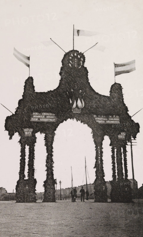 Nassauplein., Triumphal arch at Nassauplein during the April Festivals in 1887 in Amsterdam, photograph, cabinet photograph, Part of Photo album with images of the April Festivals in 1887 in Amsterdam., Amsterdam, 1887-04, photographer, pasteboard, ink, materials by function, paper, printing ink, collotype, photogravure, height 155 mm x width 95 mm, prospect of city, town panorama, silhouette of city, gate, entrance, April Festivals 1887, Nassauplein, A tall ornamental archway covered in dense foliage with multiple vertical pillars and rounded openings stands in the foreground, topped by a central rounded mound with small decorative elements and several flagpoles bearing flags; silhouettes of a few people are visible walking beneath the arch, and a row of thin masts and lamp posts can be seen in the background along a flat open area.