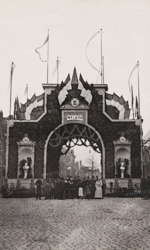 Westerstraat., Triumphal arch in the Westerstraat during the April Festivals in 1887 in Amsterdam, photograph, cabinet photograph, Part of Photo album with images of the April Festivals in 1887 in Amsterdam., Amsterdam, 1887-04, photographer, pasteboard, ink, materials by function, paper, printing ink, collotype, photogravure, height 147 mm x width 100 mm, prospect of city, town panorama, silhouette of city, gate, entrance, April Festivals 1887, Westerstraat (Amsterdam), A large ornate archway decorated with garlands and surrounded by stylized triangular pennants fills the center of the image, topped with tall poles bearing long flowing flags and streamers; two alcoves flank the arch, each containing a bust on a pedestal and framed by decorative molding, while a crowd of people stands beneath and in front of the arch on a cobbled street, with buildings and bare trees visible through the opening in the background.