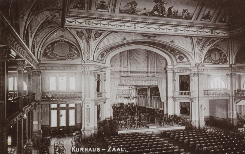 Concert hall with stage of the Kurhaus in Scheveningen, Kurhaus - Hall, photograph, cabinet photograph, Part of Leporello album with recordings of Scheveningen., Scheveningen, c. 1890 - c. 1910, photographer, pasteboard, ink, materials by function, paper, printing ink, collotype, photogravure, height 95 mm x width 147 mm, group of actors, troupe, actors on the stage, concert hall, Kurhaus (Scheveningen), A wide ornate auditorium with a large proscenium arch and heavy draped curtains framing a stage filled with stacked chairs and music stands, flanked by tall decorative columns and balconies with railings and small box seats, a painted and sculpted ceiling with decorative panels and medallions, rows of chairs arranged in the main floor seating area, arched windows along the side walls, chandeliers and decorative moldings throughout, and potted plants near the stage.