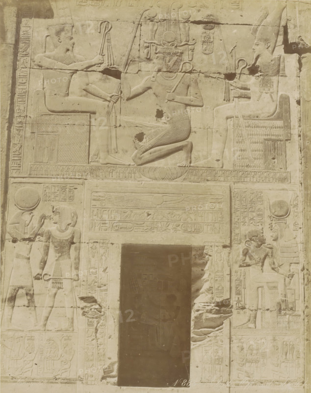 Wall with hieroglyphs and reliefs in Egypt (former title), Abydos Offrande de, Relief in the temple of Seti I at Abydos, photograph, Part of Travel album with 80 photos of Egypt., negative number: '883', photographer: Zangaki, C. & G., Egypt, c. 1870 - c. 1890, North Africa, pasteboard, paper, albumen print, height 276 mm x width 220 mm, Egyptian hieroglyphs, A stone relief panel showing carved figures arranged around a recessed dark doorway, with a central scene above the doorway of a kneeling figure facing a seated figure on the left and another seated figure on the right, each holding staffs and wearing ornate headdresses; the kneeling figure wears a pleated skirt and a tall, intricate headdress with curved elements and holds a curved staff; the seated figures wear tall crowns and collars and hold staffs and flails while seated on patterned thrones; below the central scene a horizontal band with a winged disk and carved panels extends above the doorway; flanking the doorway are vert...