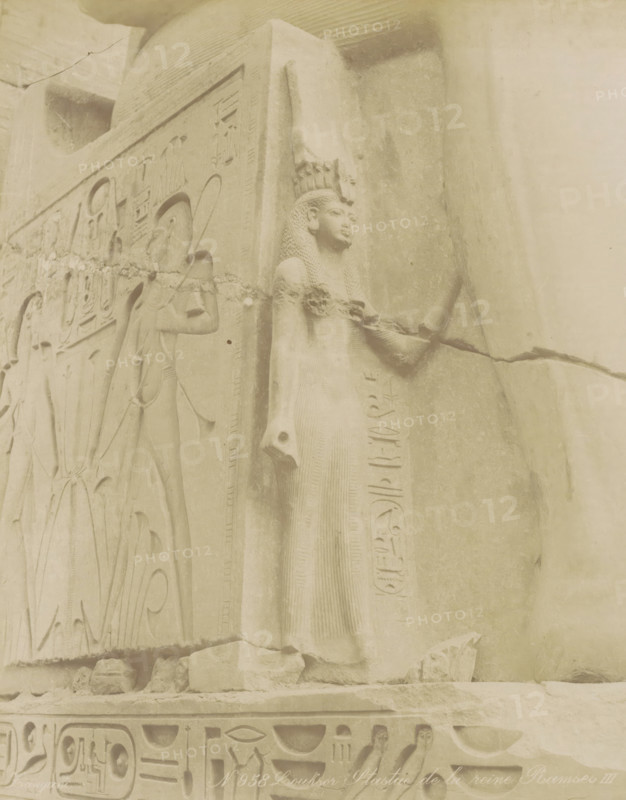 Sculpture of the queen of Pharaoh Ramses II in Luxor, Louksor Statue de la reine Ramses III, photograph, Part of Travel album with 80 photos of Egypt., negative number: '958', photographer: Zangaki, C. & G., Luxor, c. 1870 - c. 1890, North Africa, pasteboard, paper, albumen print, height 275 mm x width 220 mm, Contrary to what the title states on the object, this is Ramses II, piece of sculpture, reproduction of a piece of sculpture, A weathered stone relief showing a standing female figure in profile wearing a tall headdress and a long, textured dress, with braided hair and an arm extended forward holding an object; adjacent carved panels feature other figures in profile and decorative motifs, with bands of carved symbols along the base and side; the surface shows cracks, chips, and worn areas, and the scene is rendered in pale, sandy tones.