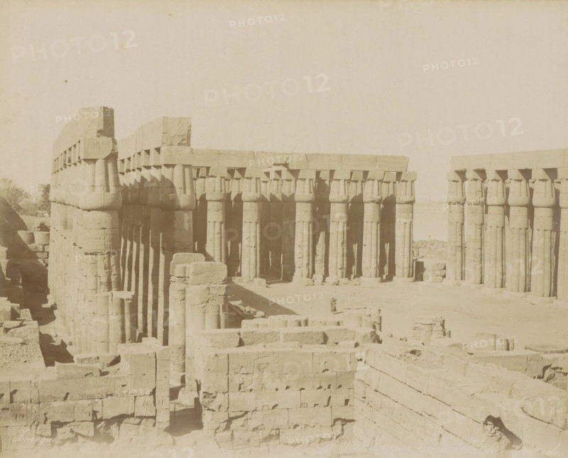 Louqsor. Ensemble de colonnade, Colonnade in Luxor, photograph, Part of Travel album with 80 photos of Egypt., negative number: '980', photographer: Zangaki, C. & G., Luxor, c. 1870 - c. 1890, North Africa, pasteboard, paper, albumen print, height 220 mm x width 276 mm, colonnade, A sepia-toned photograph of an ancient stone structure composed of rows of large vertical columns topped by rectangular beams, with some columns showing blocky capitals and horizontal stone lintels; the stone surfaces display weathering and chips, and scattered carved blocks and rubble lie in the foreground and between the columned aisles; shadows fall along the columns creating vertical dark bands, and a low horizon and pale sky fill the upper part of the image.