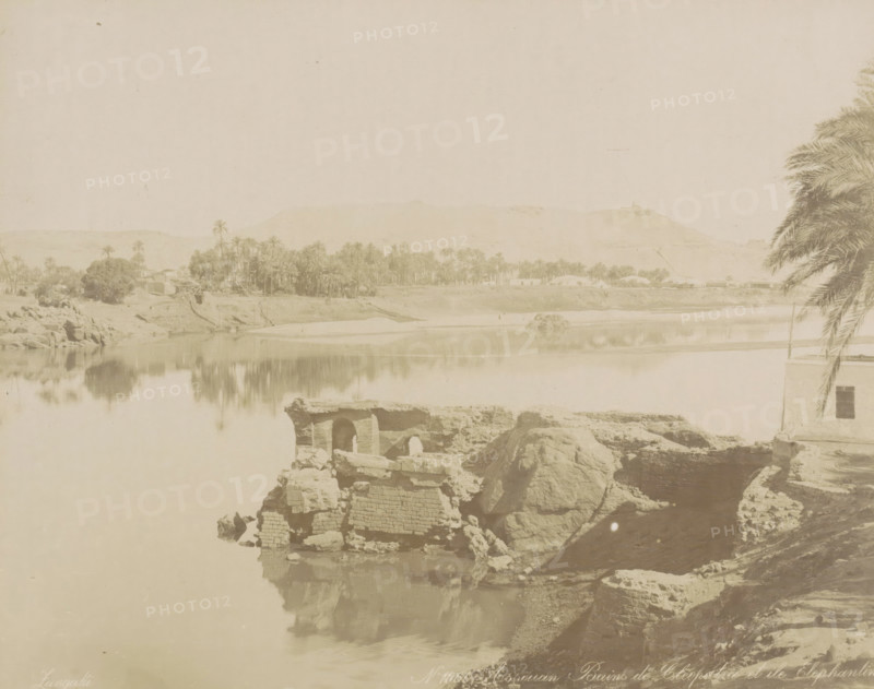 Assouan Bains de Cleopatra et ile Elephantine, Bath of Cleoptra and the island of Elephantine near Aswan, photograph, Part of Travel album with 80 photos of Egypt., negative number: '1060', photographer: Zangaki, C. & G., Aswan, c. 1870 - c. 1890, North Africa, pasteboard, paper, albumen print, height 218 mm x width 276 mm, island, river, Elephantine, A pale riverside scene showing a calm body of water reflecting the opposite bank, with clusters of palm trees and low buildings along the far shore, a range of low hills in the background, rocky ruins and large boulders in the foreground by the water's edge, and a partial view of a building with a window and an overhanging palm frond on the right side.