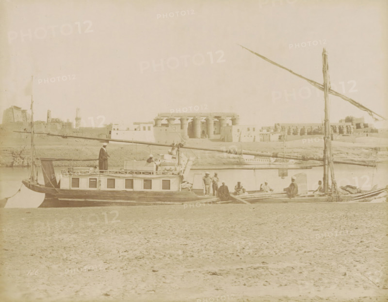 Sailboat (dahabie) on the Nile with the Great Colonnade in Luxor in the background, photograph, Part of Travel album with 80 photos of Egypt., negative number: '126', photographer: attributed to Zangaki, C. & G., Luxor, photographer: attributed to Maison Bonfils, c. 1870 - c. 1890, Attributed to, pasteboard, paper, albumen print, height 220 mm x width 283 mm, Photos in this album are by the photographers Bonfils or by C. & G. Zangaki. Therefore, the photo has been attributed to them., sailing-ship, sailing-boat, river, temple, shrine, Egyptian religion, North Africa, Nile, A sepia-toned riverside scene showing a long wooden boat with a cabin and shutters along its side, a tall mast and horizontal spars, and several people on and around the boat wearing loose garments and head coverings; in the background across the water there are large stone buildings with columns and flat roofs and a low shoreline, and in the foreground a sandy, textured riverbank occupies the lower part of the image...