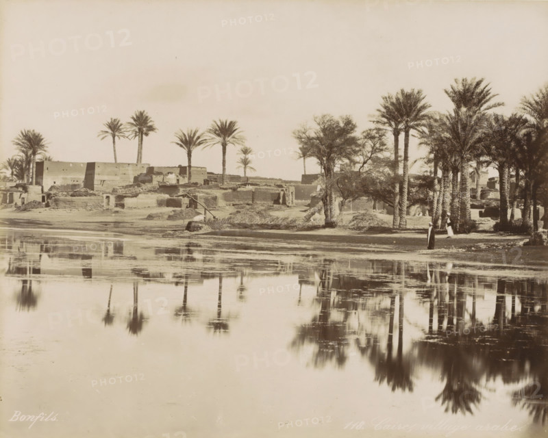 Village with palm trees by a body of water near Cairo, Caire, village arabe, photograph, Part of Travel Album with 80 Photos of Egypt., negative number: '118', photographer: Maison Bonfils, Cairo (city), c. 1870 - c. 1898, Ottoman Empire, North Africa, pasteboard, paper, albumen print, height 223 mm x width 280 mm, prospect of village, silhouette of village, trees: palm-tree, A wide sepia-toned riverside scene shows a shoreline with mud-brick buildings and low ruined structures partially obscured by clumps of vegetation. Tall palm trees rise along the bank, some clustered and some standing alone, their fronds splayed at the tops. A pair of figures in long robes stand near the trees by the water's edge. Reflections of the trees and shoreline appear in the calm river in the foreground, creating mirrored shapes and soft ripples. The sky above is pale and empty, occupying the upper portion of the image.