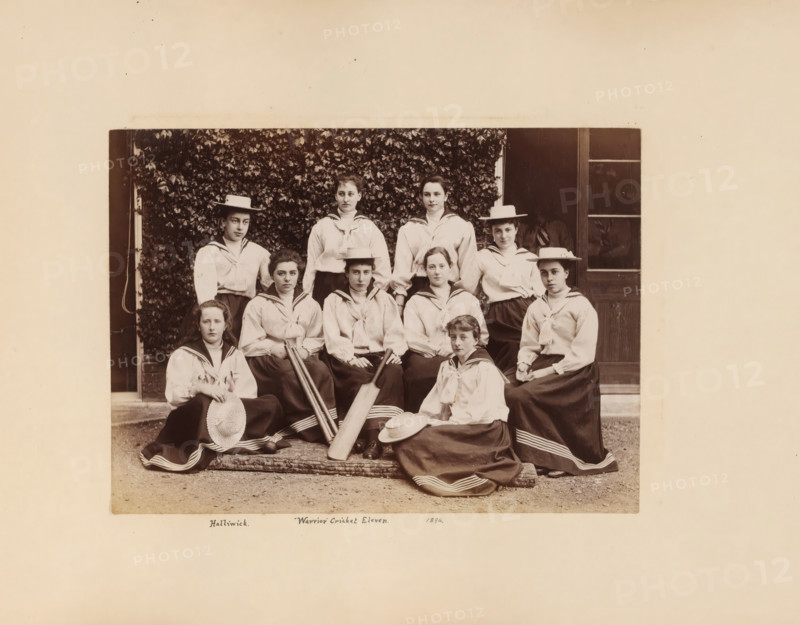 Group portrait with ten young women from the cricket team of the Halliwick School for Girls, album page, photograph, This photo is part of an album., annotation: 'Halliwick. Warrior Cricket Eleven. 1894.', London, 1894 - 1896, photographer, paper, pasteboard, albumen print, height 265 mm, width 350 mm, anonymous historical persons portrayed in a group, in a group-portrait - BB - woman, cricket (ball game), A sepia photograph shows a group of young women arranged in two rows in front of a leafy wall and a doorway, wearing light blouses with sailor-style collars and long dark skirts with light trim at the hem, some wearing flat-brimmed straw hats and others holding hats in their laps, several holding wooden cricket bats and one holding a ball, with neutral facial expressions and varied seated and standing poses on a patterned rug and a low step.